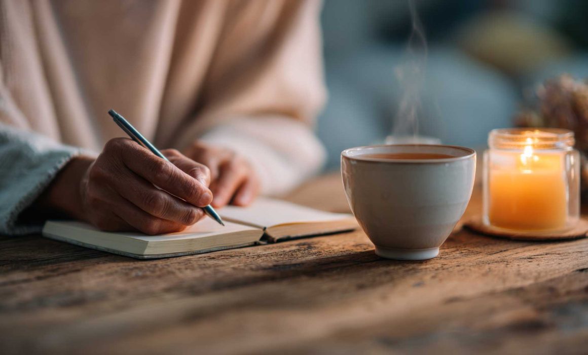 Eine Person trägt etwas in ein Buch ein, vor ihr stehen eine Kerze und eine dampfende Tasse Tee